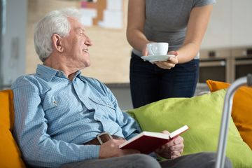 An elderly man reading an book while carer brings a cup of tea